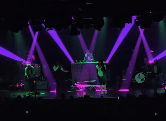 The stage during a music concert in a medium sized venue. Purple lights crisscross over a dark stage, 5 musicians are faintly illuminated by green light.