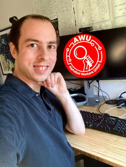 A person standing next to a computer screen, smiling. On the computer screen is a logo that says "Alphabet Workers Union"; in the background there are a series of maps.