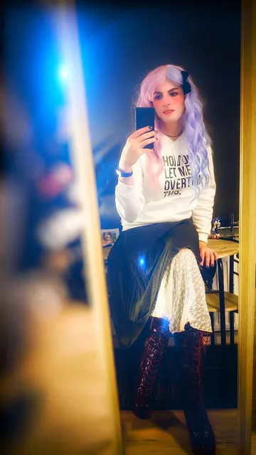 Miona taking a picture of herself in the mirror with her phone. She is sitting on a table. She is wearing a white hoodie with the text "hold on, let me overthink this.", a layered skirt with a solid dark green layer and a white mesh layer, and dark red platform boots.