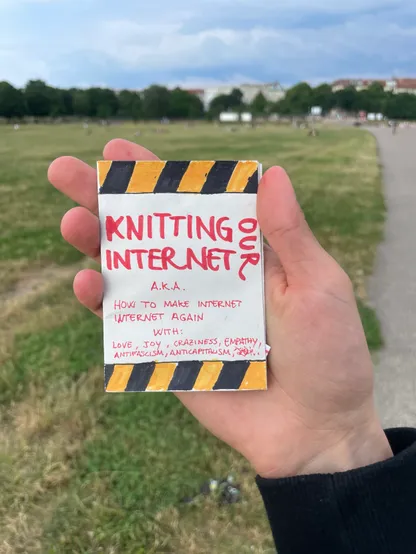 My hand holding the zine with Tempelhof Park in the background.

COVER PAGE:
A big all-caps writing with the title: “Knitting our internet”, then, written in smaller size: “a.k.a. how to make Internet Internet again – with: love, joy, craziness, empathy, antifascism, anticapitalism…”

All the writings are in red, and at the top and bottom of the page there are alternating skewed yellow and black stripes, mimicking the symbols of a work in progress.