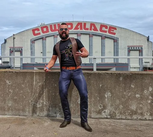 I am standing in front of the Cow Palace in San Francisco in leather jeans and a vest, with sunglasses on.