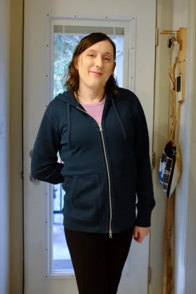 a girl standing in her doorway and smiling. she is wearing a teal hoodie and a pink top.