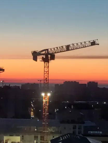 Lever de soleil pastel avec une grue en plein milieu qui se découpe et le grutier en train de monter à l'échelle