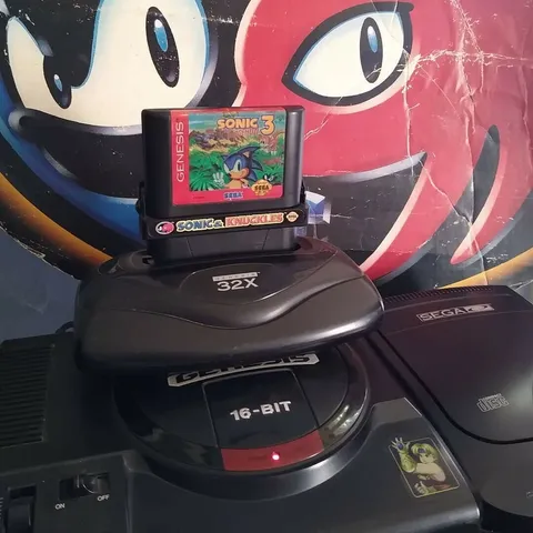 A model 1 Sega Genesis game console with a 32X and model 2 Sega CD attached to it. Otherwise known as a version of a "tower of power." A copy of Sonic and Knuckles with Sonic the Hedgehog 3 attached is inserted in the 32X. The Sonic and Knuckles game logo fills the background behind the game console.