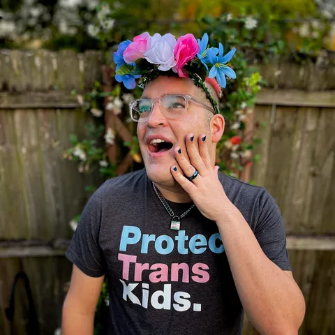 A picture of david wearing a trans flower crown and looking up to the left. They have blue painted nails with a matching blue wedding band on. They are wearing a trans flag chain collar and a shirt that says Protect Trans Kids.