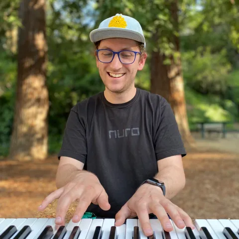 The author, wearing a black shirt, a blue flat-brimmed cap, and blue glasses, playing the keyboard in a park.