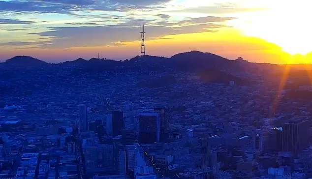 Sutro Tower in the center of the picture with mountain as a blue silhouette. Orange skies during sunset.