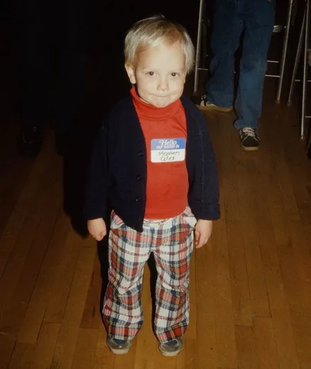 A photo of a young, blond, white boy in a red-orange turtleneck, blue blazer, and plaid pants. He is wearing a tag that says “Hello my name is (in swashy 1970s type) Stephen Coles (handwritten)”. 