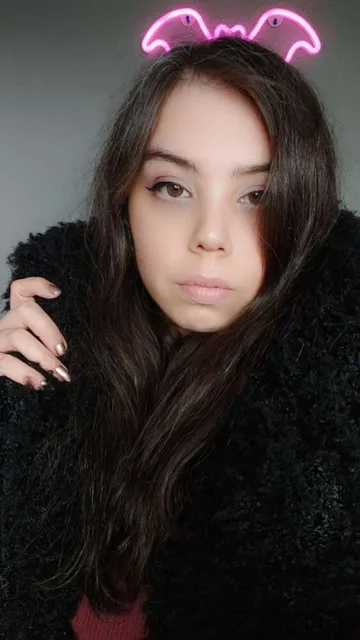 extinct irl selfie: a nonbinary femme with long dark brown hair streaked with grey, green eyes with central heterochromia, rose gold painted nails, and a black fluffy coat