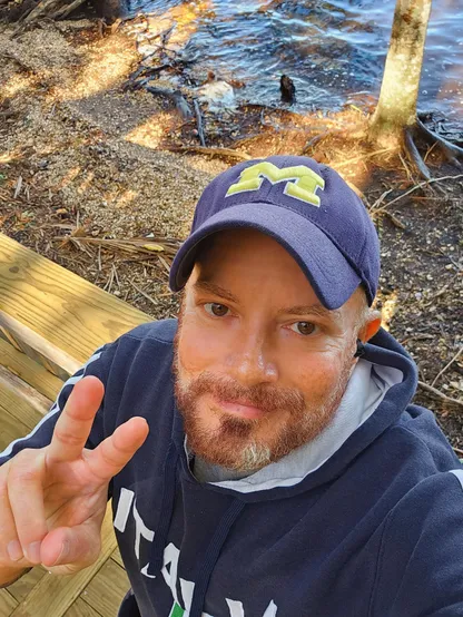 Man wearing a blue hoodie and University of Michigan ball cap, standing at the edge of a river