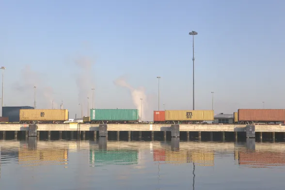 Colorful shipping containers in a line of trucking chassis on a low cement bridge over water. In the background are plumes of steam or smoke rising from petroleum refining, creating a faint brown hue that is muddying the blue sky overhead. The image is also reflecting in the water beneath the line of container. Photo: McKenzie Stribich