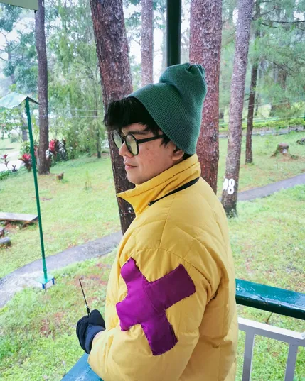 John is cosplaying KillJoy from the game Valorant. He is wearing a large yellow puffer jacket with a large print of the letter X on the sides. On his head is a green beanie and he is wearing his round glasses. He is standing on a porch looking away from the camera.