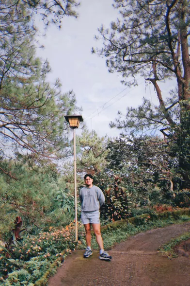 John stands next to a lamp post in a street surrounded by plants and pine trees. He is wearing a white short shorts and a large gray sweatshirt. The area is foggy and a little too cold for him to be wearing shorts.