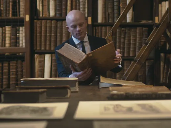 Daniel is standing in an old library, surrounded by big bound books, and  looking bemused into an old big book