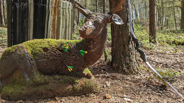 3D archery target in the shape of a sitting deer, with 3 arrows in its middle and a bow resting on a nearby tree.