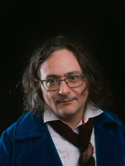 A nonbinary white person with long, wavy brown hair, green metal wayfarer glasses over hazel eyes and thick eyebrows, a prominent forehead and large Irish nose. They've a light mustache and a slight smile. They are wearing a blue velvet jacket over a button down shirt, with a scarf tied around their neck.