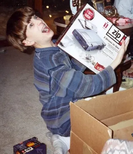 Child Nick throws his head back cackling while holding up the box of a Zip Drive that he just unwrapped for Christmas. He has a bowl cut and braces and is wearing a baggy blue shirt. A six pack of extra disks is on the floor next to the package.