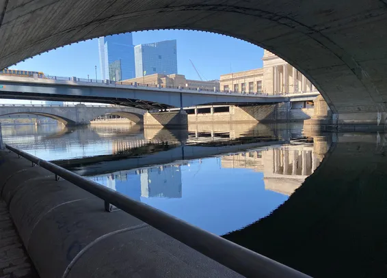 View of the Schuylkill River from the Schuylkill River Trail -- perspective is under one bridge (arching across the top of the image) and looking down river at several more.  The bridges appear in shades of tan stone, with some blue metal; the sky above and water below are blue; and there are buildings visible above the bridges -- two modern blue glass and one, to the side, with classical sandstone facade and columns.