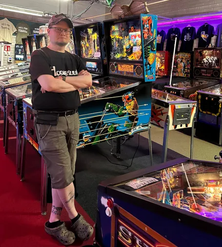 View inside an arcade full of pinball machines. A guy in a black t-shirt and baseball cap is leaning against a pinball machine.