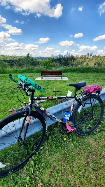 A single speed road bike in front of a bench and the Edmonton skyline in the background. The bike has a ton of stickers on it and green handlebar tape. 