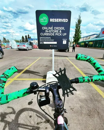 POV: you're on a road bike, looking forwards over the handlebars. They are wrapped in green bar tape, there's a black bell, a phone holder, and an action cam attached. You are in a parking spot that says RESERVED. ONLINE ORDER, IN-STORE & CURBSIDE PICK UP. For some reason, I get weird looks when I don't pull up in a huge SUV to pick up an order...