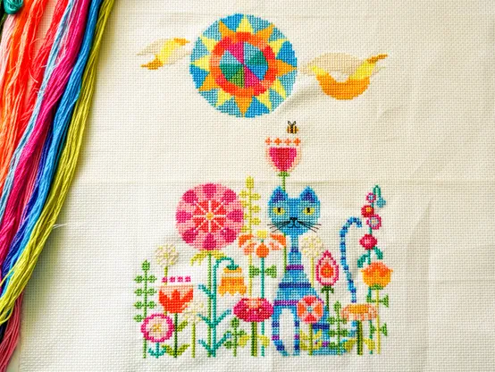 A completed cross stitch project that needs to be ironed! It's a blue cat sitting in a bright and colourful garden full of flowers.