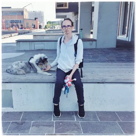 Flaki sitting on a bench in Tallinn with the coastal Noblessner area in the background. He has a white hoodie, jeans and lace-less black shoes, looking directly into the camera a bit squinty. His right hand is held up to the side, where Lumi the young blue merle collie is lying on the bench, sniffing his palm in search of treats. His left hand is full of colorful dog things, including a fox toy and Lumi's bright blue lead. The rest of the picture has a an overall faded, bluish tint and a white border that makes it almost feel like an album cover.