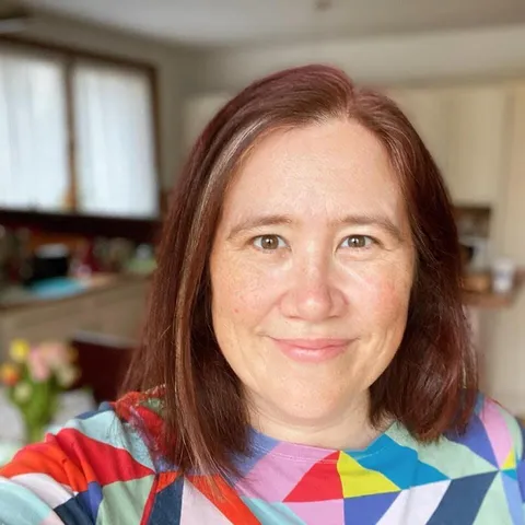 Selfie of a white woman with reddish hair (with a prominent grey streak) and freckles. She is wearing a geometric rainbow coloured t-shirt.