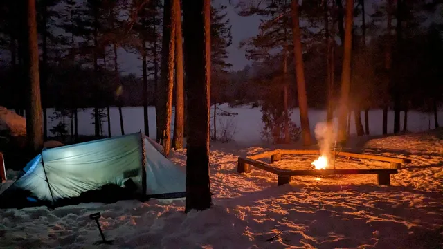 A camp in the snow amongst trees, next to a frozen lake. A green tent lit from the inside, pitched next to a campfire. The scene is coloured orange by the glow from the fire.