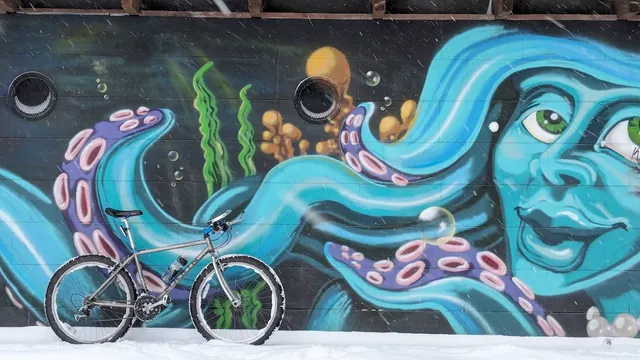 An old mountain bike leaning up against a marine themed mural. There is snow on the ground and the bike's tires are moderately caked in snow.