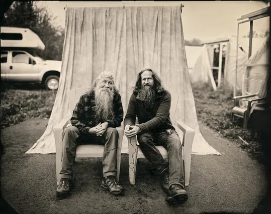 Two men in their 60s with long hair and long beards sit side by side, facing the camera while leaning toward each other. A photographic backdrop curtain hangs behind them, but they're clearly outside on a driveway. To the left, the front end of a pickup truck with an over-the-cab camper is visible. On the right are the front entry ways to a series of greenhouses.  The image itself was made using a glass negative technique from the 1800s. Not quite black and white , it's more of a light tan and black image.