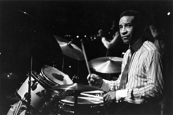 Max Roach Like No Other
Photograph by David Coleman 

(The drummer Max Roach, wearing a striped shirt, a tie, behind drums, the left hand stick on snare drum, the right hand stick raised to ride cymbal)