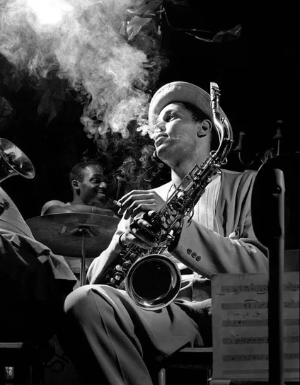 Dexter Gordon, dressed with costume and hat, in a cloud of cigarette smoke, saxophone on the lap, a music sheet on the right. Black and white photograph by Herman Leonard, 1948