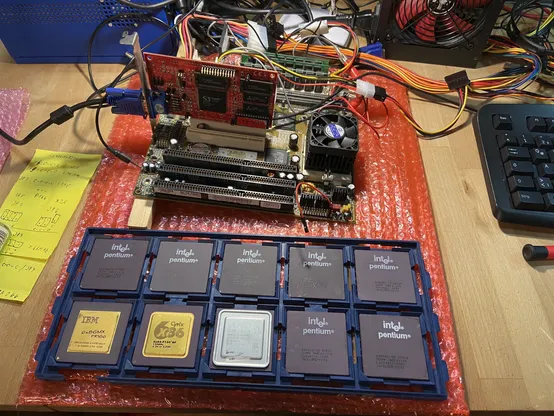 a pc mainboard with cards on a table, wired to a power supply with a tray of CPUs in front
