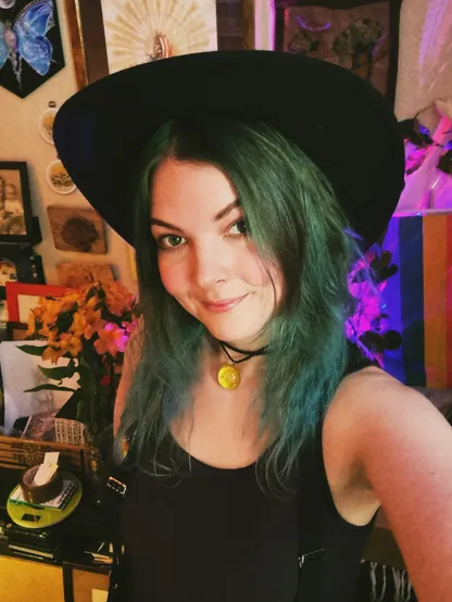 Selfie - Jen's wearing a black hat. She has greenish hair and is wearing a tanktop with suspenders. There's a bunch of pictures up on the wall behind her, along with a rainbow flag. She looks pretty content in general.