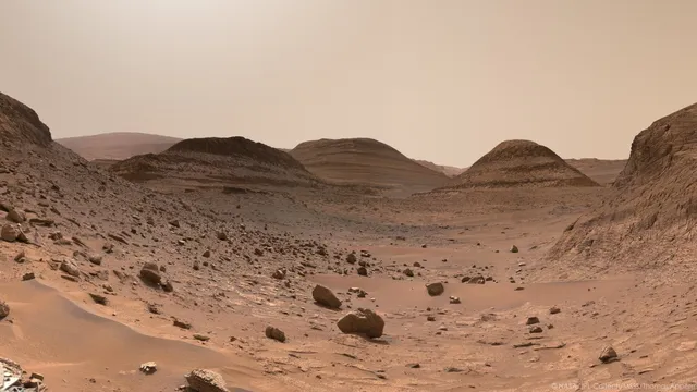 A picture of Mars taken by Curiosity rover. It shows a valley bordered by mounds and stratified, in a region where there was once liquid water. Sulfates salts have been deposited there. We see a nice little dune and rocks in the foreground, fallen from the top of the mounds. In the background, we see Mount Sharp, a 5500 meters high mountain at the center of Gale crater.