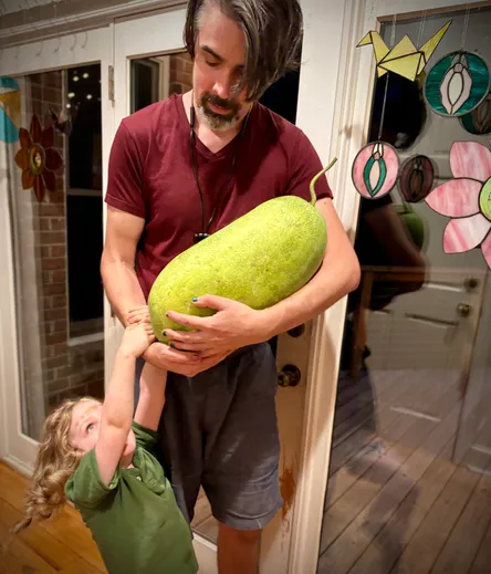 An adult, cradling a large watermelon like a baby, while a kid hangs on his arm. Stained glass paper cranes and vulvas and flowers hanging in the window.