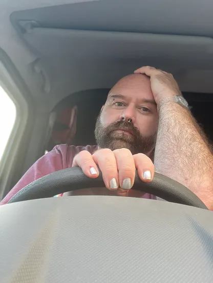 Image shoes Rob sitting in the driving seat of a minibus. He’s parked up waiting for his boss. His nails are painted silver and he has one hand on the steering wheel and another on his head. He doesn’t look happy. 