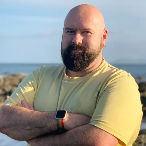 Image shows Rob stood by the coast. He is wearing a yellow tee and his arms are folded. 