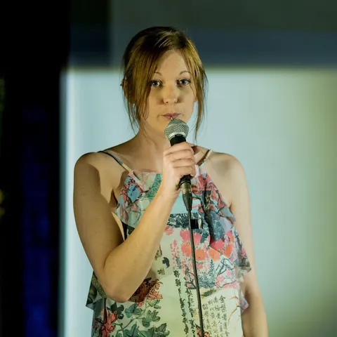 A woman with dark hair and a lip piercing, wearing a patterned pale green top, on stage holding a microphone and making eye-contact with the camera. 