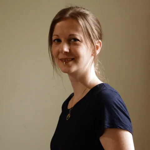 A woman with a lip piercing and medium-brown hair tied back, wearing a dark blue t-shirt and a silver necklace. 