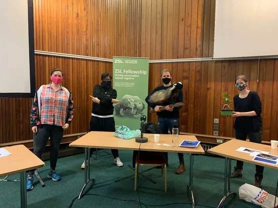 Claire Asher, Billy Heaney, Sue Perkins and Josie Long wearing face masks and standing behind three desks with microphones on them at the Nature Table recording at London Zoo. Billy is holding a stuffed badger, Claire is holding a bonsai tree. 