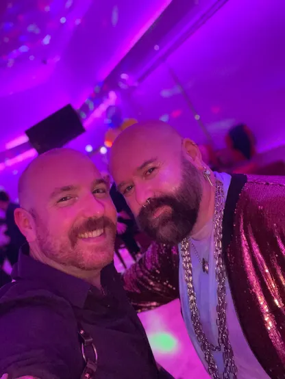 Me and my husband at a “glitter and gold” themed party. We’re smiling at the camera, wearing golden accessories and the lighting behind us is purple. 