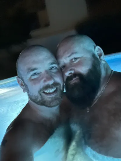 A selfie of me and my husband stood in a swimming pool at night. We’re visible from the chest up, standing close together, cheek to cheek. The background is dark and our faces are lit with a blue glow by the lights under the water.