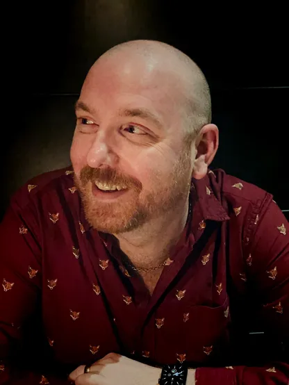 Me sat in a restaurant, looking off to the left of the camera. The lighting is dim, the background is dark. I’m smiling. 