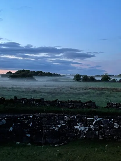 A view across the fields in Autumn. It’s twilight, there’s a stone wall in the foreground and there’s mist obscuring the fields. 