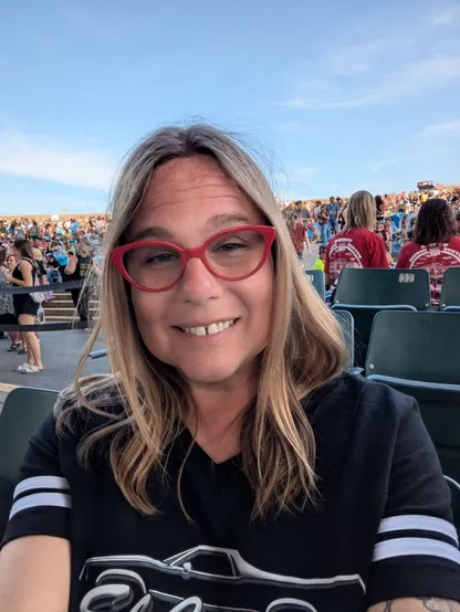 Selfie of Kit - a woman with frazzled blonde hair, cat-eye glasses with red frames, wearing an El Camino t-shirt. Behind her is a crowd of people in a music stadium.