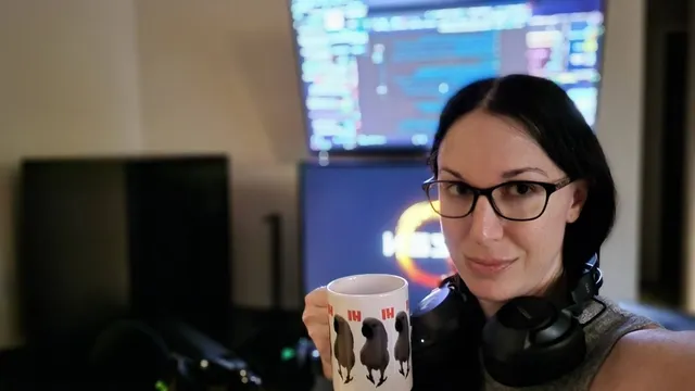 A selfie of a woman with black hair and glasses holding a coffee mug in front of a computer setup. 