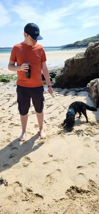 Middle aged white man in blue cargo shorts and a red t-shirt, baseball cap standing barefoot on a sunny Cornish beach. He is looking down at a black and working cocker spaniel cross dog.