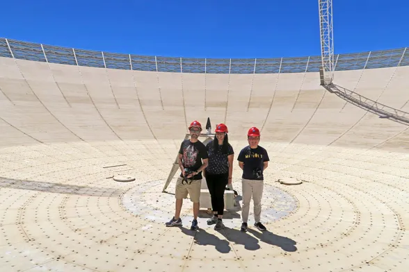 three people, wearing hard hats and standing in the middle of a large, radio telescope dish antenna